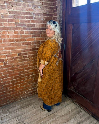 A woman with blonde hair stands in a rustic room with a brick wall and a dark wooden door, wearing a mustard yellow patterned long-sleeve tunic dress with blue jeans.