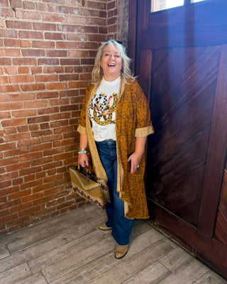 A woman stands in a rustic room with a brick wall and wooden door, wearing a mustard yellow patterned cardigan over a white graphic t-shirt, blue jeans, and tan boots, while holding a brown suitcase.