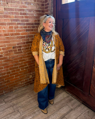 A woman stands in a rustic room with a brick wall and dark wooden door, wearing a mustard yellow patterned cardigan over a white graphic tee, blue jeans, and tan boots.