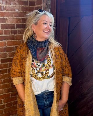A woman stands in a rustic room with a brick wall and dark wooden door, wearing a mustard yellow patterned cardigan over a white graphic tee, blue jeans, and tan boots.
