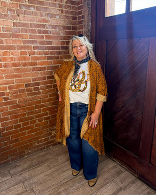 A woman stands in a rustic room corner with a red brick wall and a dark wooden door, wearing a mustard yellow patterned cardigan over a white graphic t-shirt, blue jeans, and brown shoes.