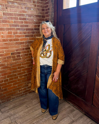A woman stands in a rustic room corner with a red brick wall and a dark wooden door, wearing a mustard yellow patterned cardigan over a white graphic t-shirt, blue jeans, and brown shoes.