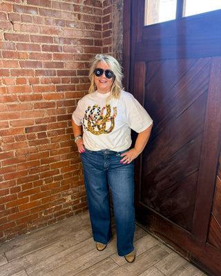 A woman wearing a white graphic t-shirt with a gold and multicolor anchor design, blue jeans, and sunglasses, standing in a corner with a brick wall, wooden door, and wood floor.