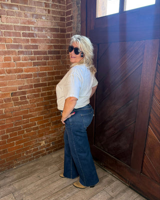 A woman with blonde hair wearing a white short-sleeve top, dark blue flared jeans, and sunglasses stands in a corner with a brick wall, wooden door, and wood floor.