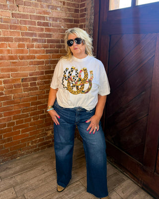 A woman with blonde hair and sunglasses stands in a brick and wood doorway wearing a white graphic t-shirt with a gold and multicolor horseshoe design, paired with blue jeans.