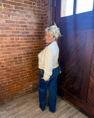 A woman with blonde hair stands in a corner of a room with a red brick wall, a dark wooden door, and wood-look flooring, wearing a cream long-sleeve top and dark blue wide-leg jeans.