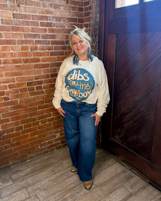 A woman stands in a rustic indoor corner with a red brick wall and dark wooden door, wearing a cream sweatshirt with a blue circular graphic that reads 'dibs on the cowboy' and blue jeans.