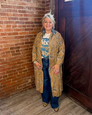 A woman stands in a rustic indoor setting with a red brick wall, wooden door, and wood floor, wearing a long tan paisley cardigan over a graphic t-shirt and blue jeans.