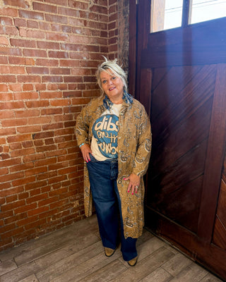 A woman stands in a rustic room with a brick wall, wooden door, and wood floor, wearing a long beige paisley cardigan over a graphic t-shirt, blue jeans, and brown shoes.
