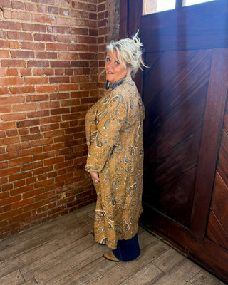 A woman with short blonde hair stands in a corner of a room with a red brick wall, dark wood door, and wood floor, wearing a long tan patterned coat over a dark blue top and dark pants.