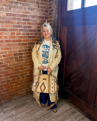 A woman stands in a rustic room with a brick wall and wooden door, holding a tan woven handbag with geometric patterns. She is wearing a beige cardigan over a graphic t-shirt.