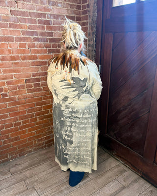 A person viewed from behind wearing a long, distressed beige coat with dark graphic text and illustration, standing in a corner with a red brick wall, dark wood door, and wood floor.