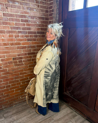 A woman with short blonde hair stands in a corner of a building with a red brick wall, dark wood door, and wood floor, wearing a long beige trench coat over a dark patterned dress or outfit with blue boots and carrying a tan shoulder bag.