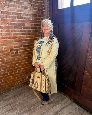 A woman stands in a rustic room with a brick wall and wooden door, wearing a cream-colored coat and holding a beige tote bag with geometric patterns.