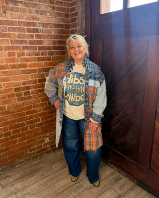 A woman stands in a rustic indoor setting with a brick wall and a dark wooden door, wearing a layered outfit consisting of a beige graphic t-shirt under a plaid overshirt and a denim jacket.