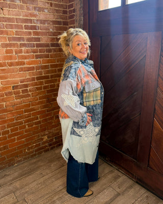 A woman stands in a rustic room with a brick wall and dark wooden door, wearing a long patchwork cardigan made from denim, plaid, lace, and light blue fabric, paired with dark jeans.