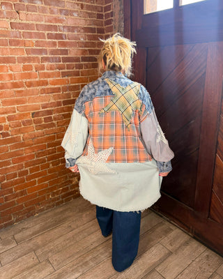A person viewed from behind wearing a patchwork quilted jacket with a plaid pattern, star applique, and lace sleeves, standing in a room with a brick wall and wooden door.
