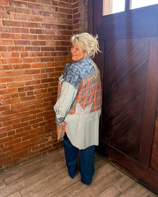 A woman with blonde hair stands in a rustic room with a brick wall and wooden door, wearing a patchwork quilted jacket with blue, orange, and beige panels, dark blue jeans, and black boots.
