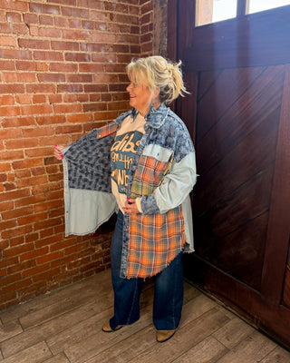 A woman stands in a room with a brick wall and wooden door, wearing a multi-panel patchwork denim jacket over a graphic tee and jeans.