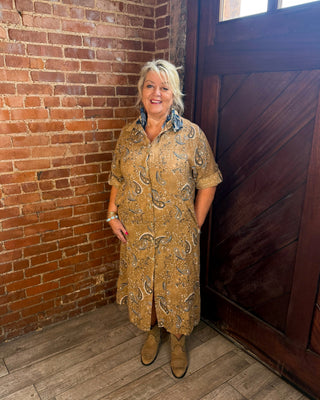A woman stands in a rustic room with a red brick wall and a dark wooden door, wearing a tan and brown paisley-patterned shirt dress with a blue patterned collar and tan boots.