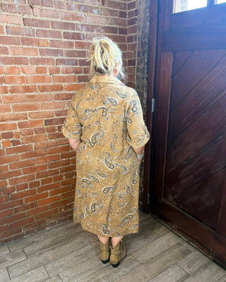 A woman stands with her back to the camera wearing a tan and brown paisley patterned cardigan or coat, standing in a corner with a red brick wall, dark wood door, and wood floor.