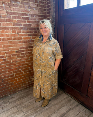 A woman stands in a rustic room with a brick wall and wooden door, wearing a tan and brown paisley patterned long-sleeve shirt dress with brown boots.
