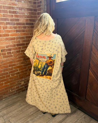 A woman stands with her back to the camera in a rustic room with a brick wall and wooden door, wearing a cream-colored dress with a small repeating pattern and a large vintage-style 'Texas' graphic print on the back.