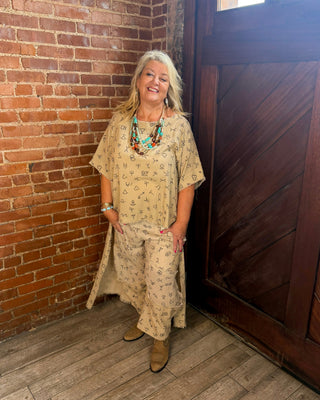 A smiling woman with blonde hair stands in a rustic room with a brick wall and a large wooden door, wearing a beige patterned tunic dress with matching pants and brown boots.