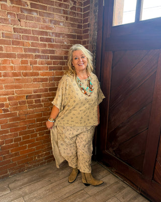A woman with blonde hair stands in a rustic room corner with a brick wall, wooden door, and wood floor, wearing a beige patterned tunic and pants set with brown boots and turquoise jewelry.