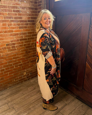 A woman stands in a rustic indoor setting with a brick wall, wooden door, and wood floor, wearing a long cream apron-style top over a black floral kimono-style dress with orange and brown accents, paired with tan boots.