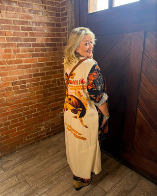 A woman stands in a corner with a brick wall and dark wooden door, wearing a long cream vest with a large brown and orange graphic print on the back, layered over a dark patterned top.