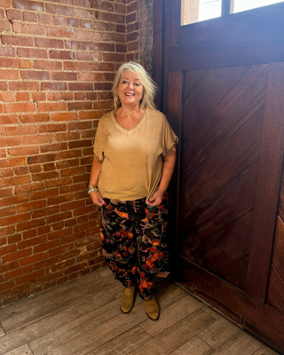 A woman with blonde hair stands in a corner of a room with a red brick wall, dark wood door, and wood floor. She is wearing a beige short-sleeve top, black pants with a red and orange floral pattern, and tan boots.