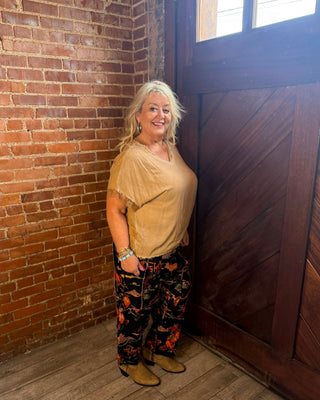 A woman with blonde hair stands in a rustic room corner with a brick wall, wooden door, and wood floor. She is wearing a beige short-sleeve top, black pants with a red and orange floral pattern, and tan boots.