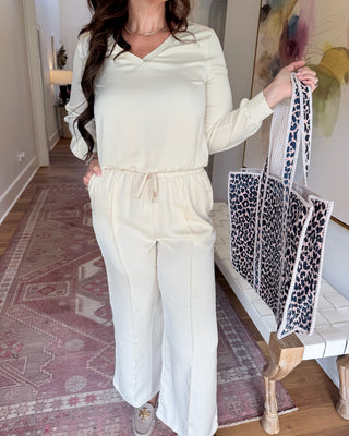 A woman wearing a cream-colored long-sleeve V-neck jumpsuit with a drawstring waist, holding a black and white leopard print tote bag in a home hallway.