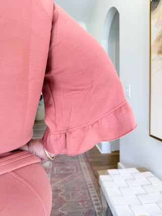 A close-up of a person wearing a dusty pink matching set, featuring a textured top with wide flared bell sleeves that have ruffled trim, paired with matching pink bottoms. The background shows a home interior with a patterned rug, white wall, and a woven bench.