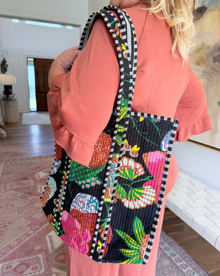 A person wearing a coral pink outfit carries a large quilted shoulder bag with a colorful floral and butterfly pattern on a black background, set in a bright indoor room.