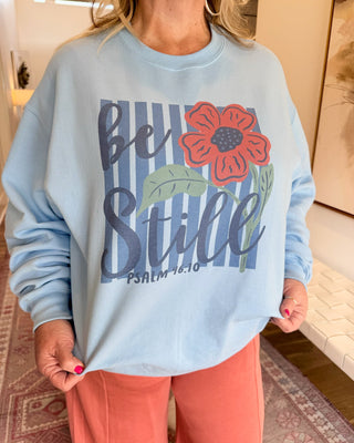 A person wearing a light blue crewneck sweatshirt with a graphic that reads 'be Still Psalm 14:10' over blue vertical stripes, alongside a red flower and green leaves, paired with coral pants in an indoor setting.