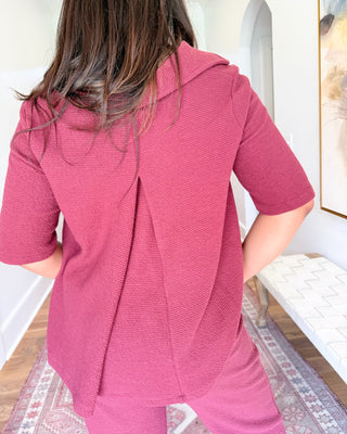 A woman shown from behind wearing a textured pink short-sleeve top and matching pants in a bright indoor room with white walls and a patterned rug.
