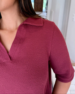 A woman with long dark brown hair smiling and looking to the side, wearing a textured short-sleeve polo shirt in a deep berry or burgundy shade, set against a bright, modern home interior background.