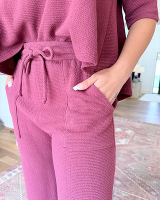A person wearing a matching textured waffle-knit top and pants set in a dusty rose or mauve pink color, standing indoors with one hand in a pocket.