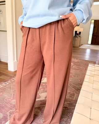 A person wearing dusty rose wide-leg pleated pants and a light blue sweatshirt, standing in a home interior with wood floors and a patterned rug.