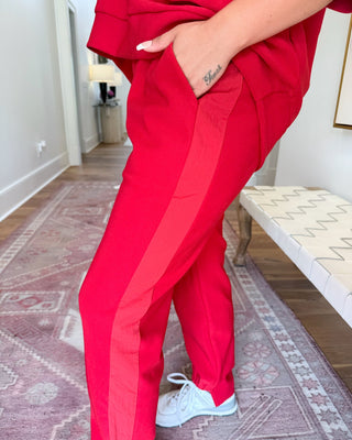 A person wearing bright red scrub-style pants with a lighter red side stripe, standing indoors on a patterned rug near a bench.
