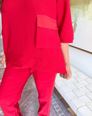 A person wearing a bright red matching lounge set with a short-sleeve top featuring a large patch pocket and coordinating pants, shown in an indoor setting with a white woven bench and patterned rug.