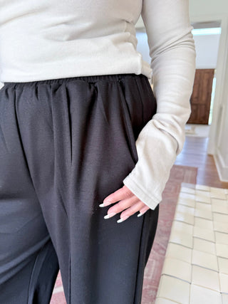 A person wearing black wide-leg lounge pants and a cream long-sleeve top, standing in a tiled indoor hallway.