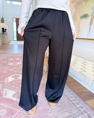 A person wearing loose black wide-leg pants with a drawstring waist, paired with a white long-sleeve top and tan shoes, standing on a patterned rug in a home interior.