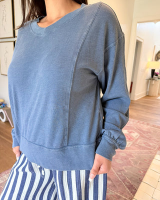 A person wearing a dusty blue long-sleeve sweatshirt and blue-and-white striped pants, standing in a home interior with wood floors and a patterned rug.