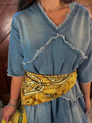 A person wearing a light blue denim dress with frayed trim and a wide mustard yellow patterned sash tied at the waist, photographed against a wooden wall.