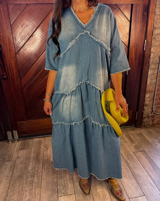 A woman wearing a light blue denim tiered maxi dress with raw-edge detailing, holding a bright yellow hat, standing in front of a rustic wooden door.