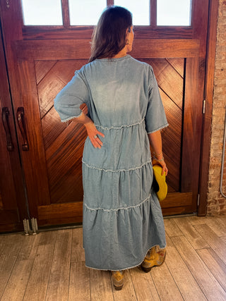 A woman stands with her back to the camera wearing a light blue tiered denim maxi dress, holding a yellow hat in a rustic room with a wooden door and brick wall.