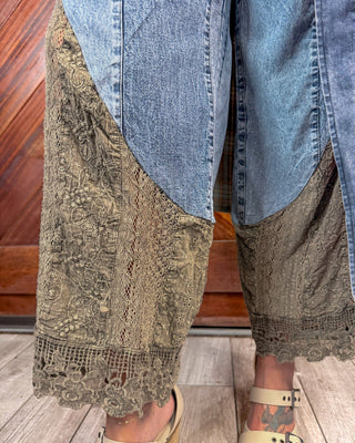 Close-up of a person's legs wearing wide-leg denim pants with large olive-brown lace overlay panels and beige sandals, standing on a wood floor.